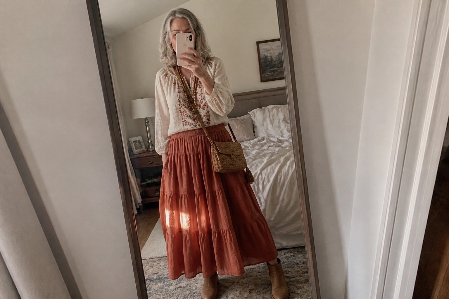 Silver-haired woman in embroidered cream blouse and tiered rust skirt taking mirror selfie.