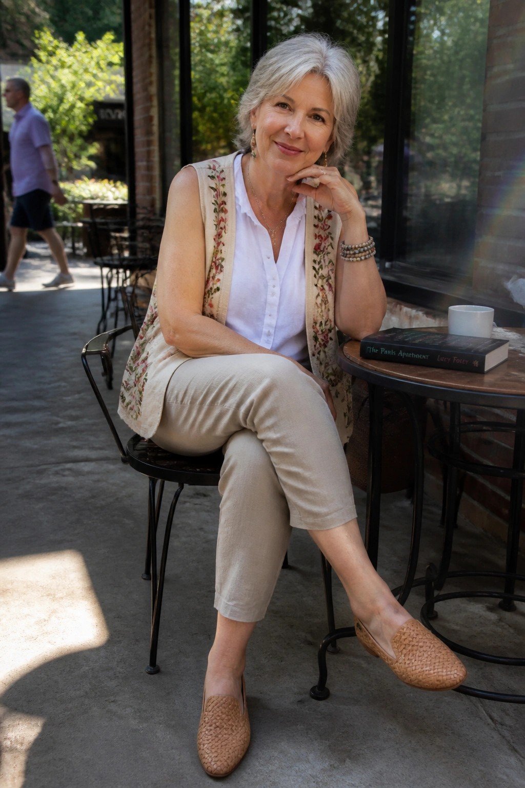 Woman over 60 wearing a white sleeveless button-up shirt layered under a beige floral embroidered vest, paired with cropped beige pants and woven beige loafers, accessorized with gold jewelry and bracelets.