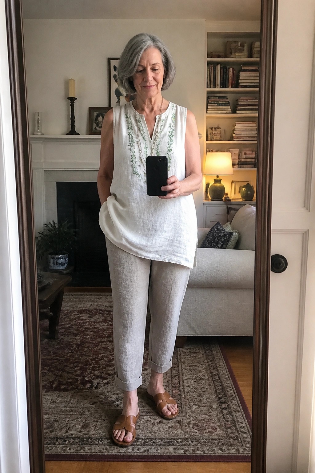 Woman over 60 in white embroidered linen tunic, light neutral linen pants cuffed at the ankle, and brown leather slide sandals holding a phone.