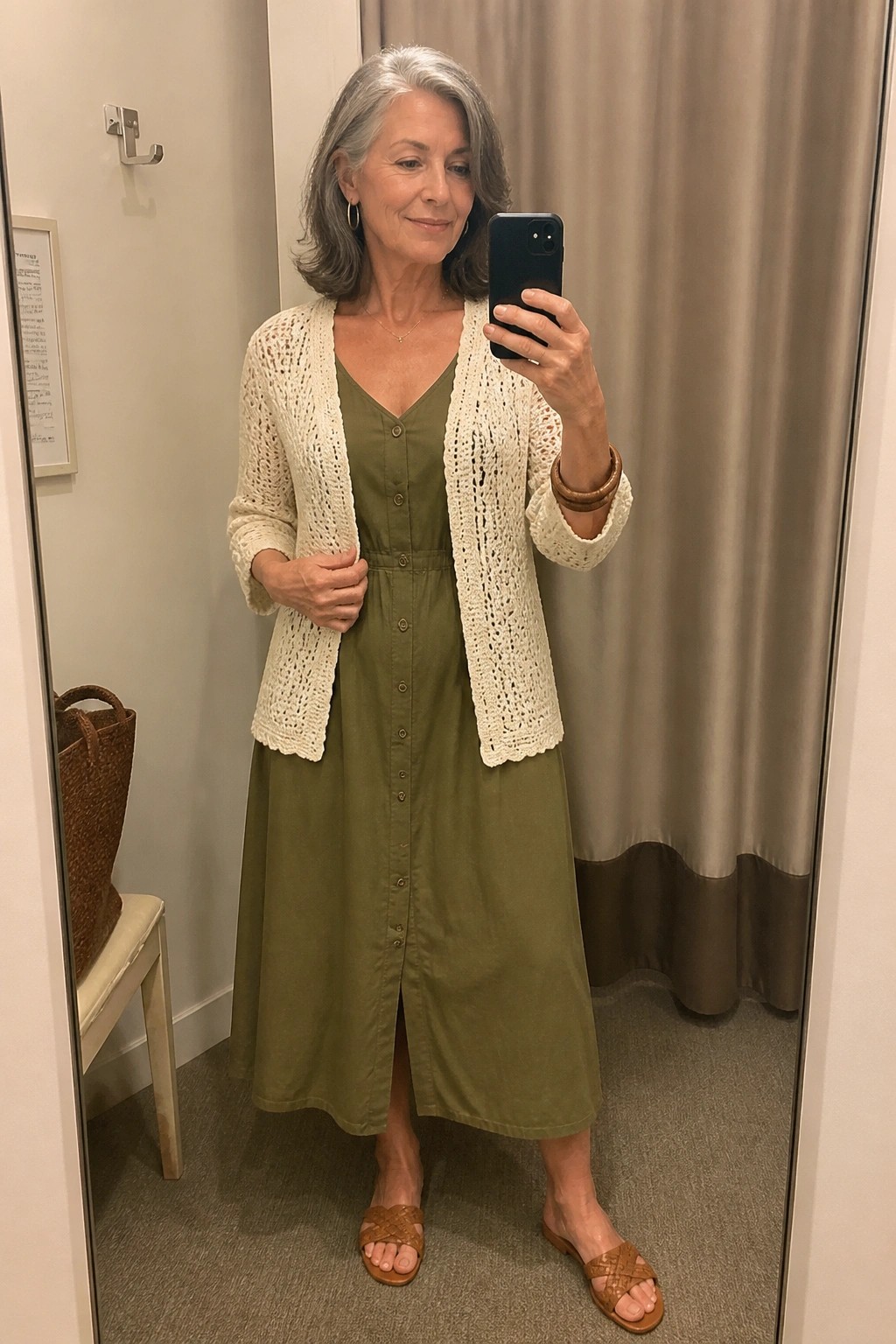 Gray-haired woman in green midi dress and cream crochet cardigan taking mirror selfie.