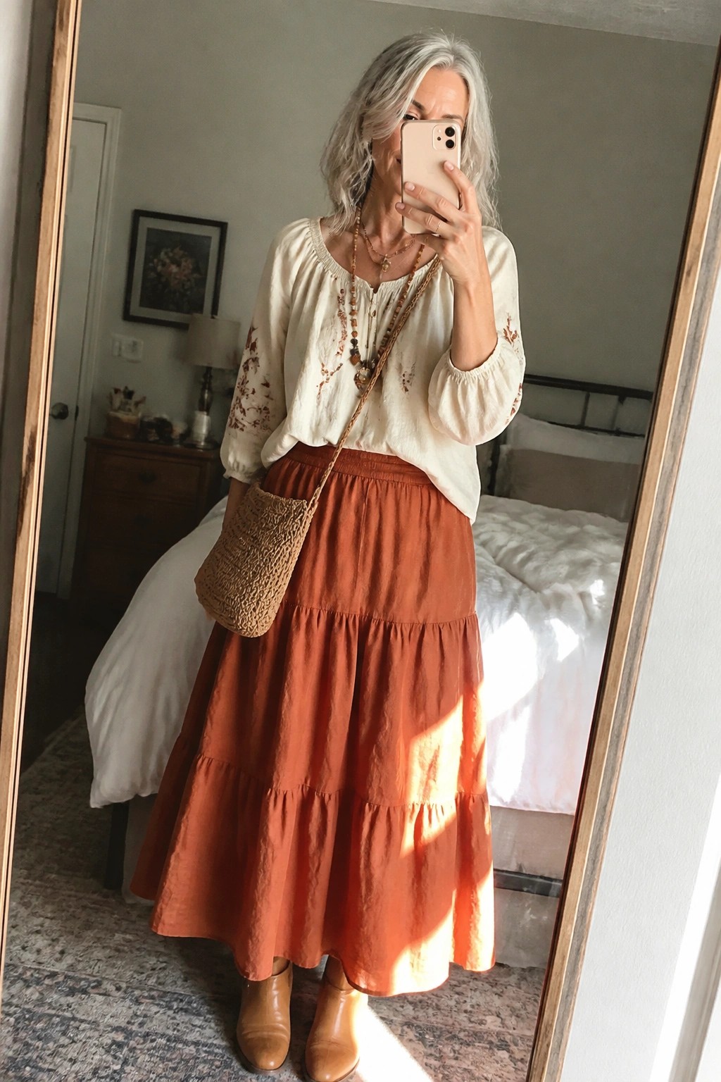 Silver-haired woman in embroidered cream blouse and tiered rust skirt taking mirror selfie.
