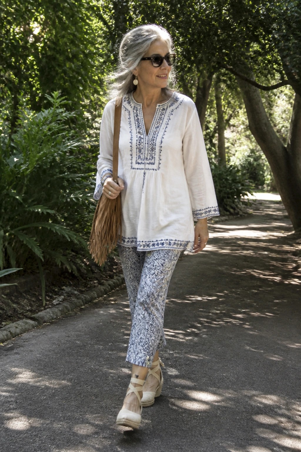 Woman over 60 wearing a white embroidered tunic top with blue details paired with blue and white patterned cropped pants, a fringed brown bag, and neutral wedge espadrilles on a park path.