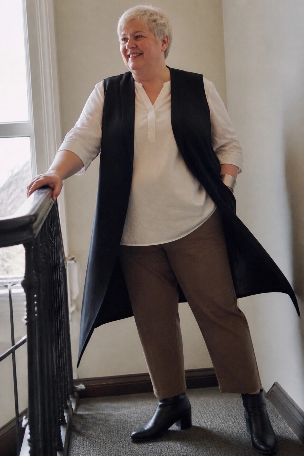 Smiling short-haired woman in long black vest over white blouse and brown pants on staircase