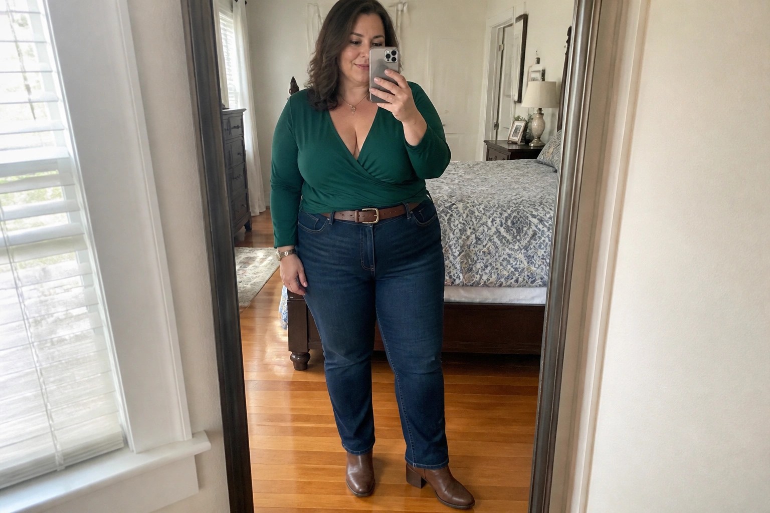 Woman in green wrap top and blue jeans taking mirror selfie in bedroom