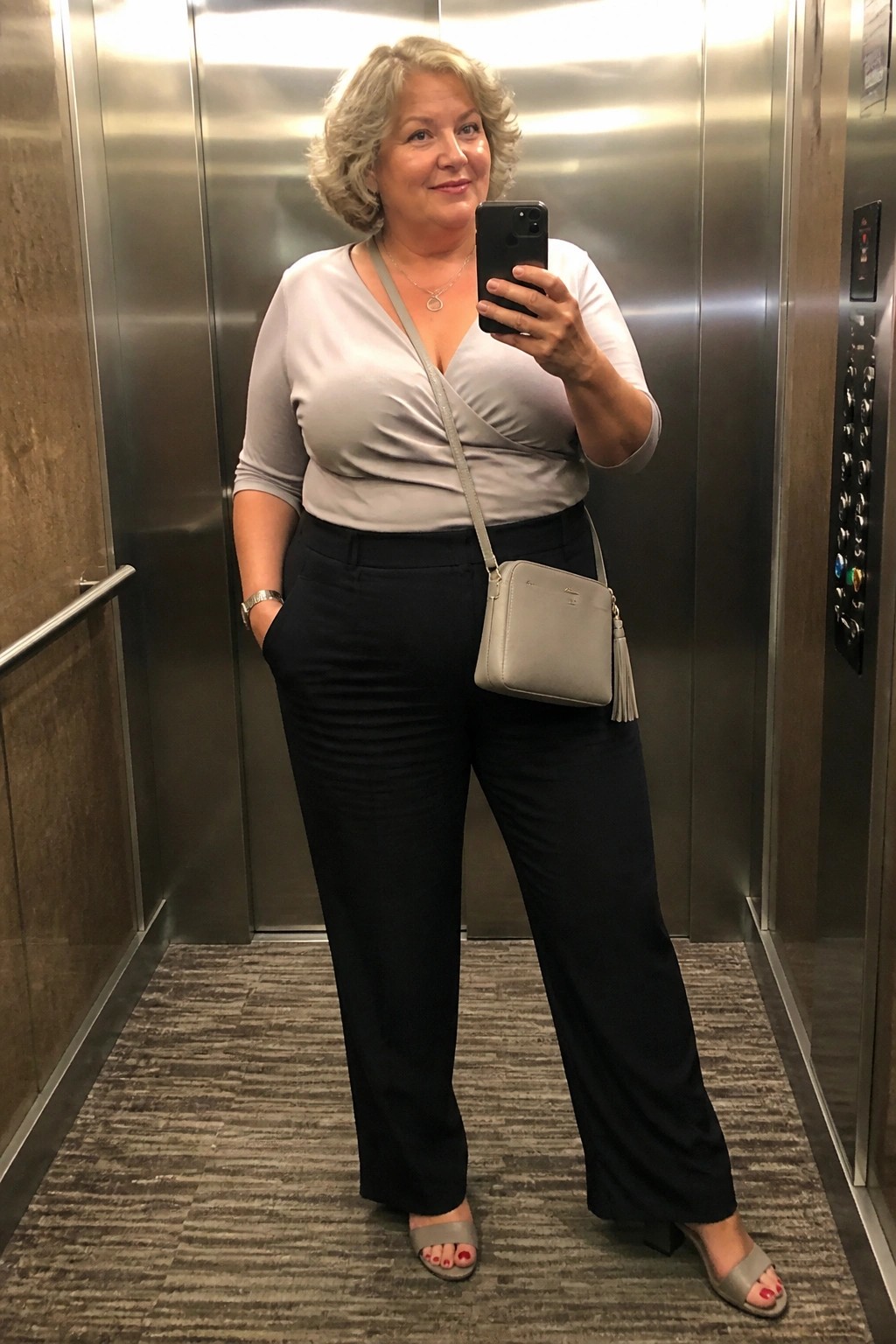 Woman taking selfie in elevator wearing gray top, black pants, and gray purse