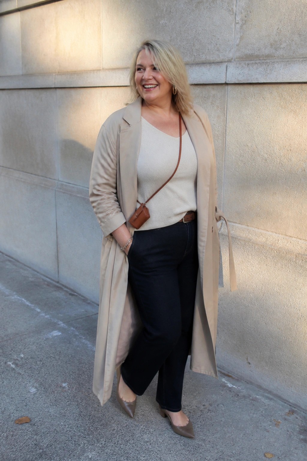 Smiling blonde woman in beige trench coat, cream sweater, and black jeans outdoors