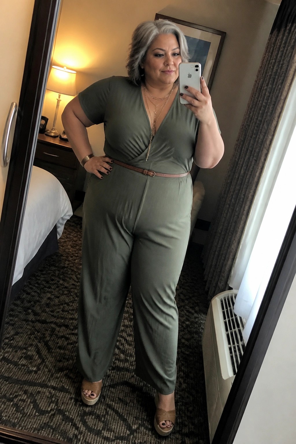 Woman with gray hair in green jumpsuit taking mirror selfie in hotel room