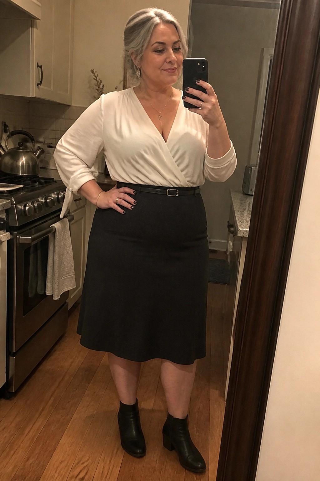 Woman over 50 wearing a white wrap blouse tucked into a black midi skirt with a thin black belt and black ankle boots while taking a mirror selfie in a kitchen.
