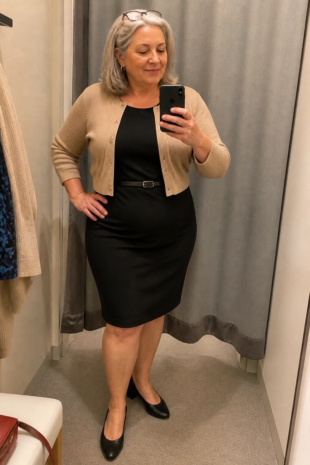 Woman with gray hair in black dress and beige cardigan taking mirror selfie