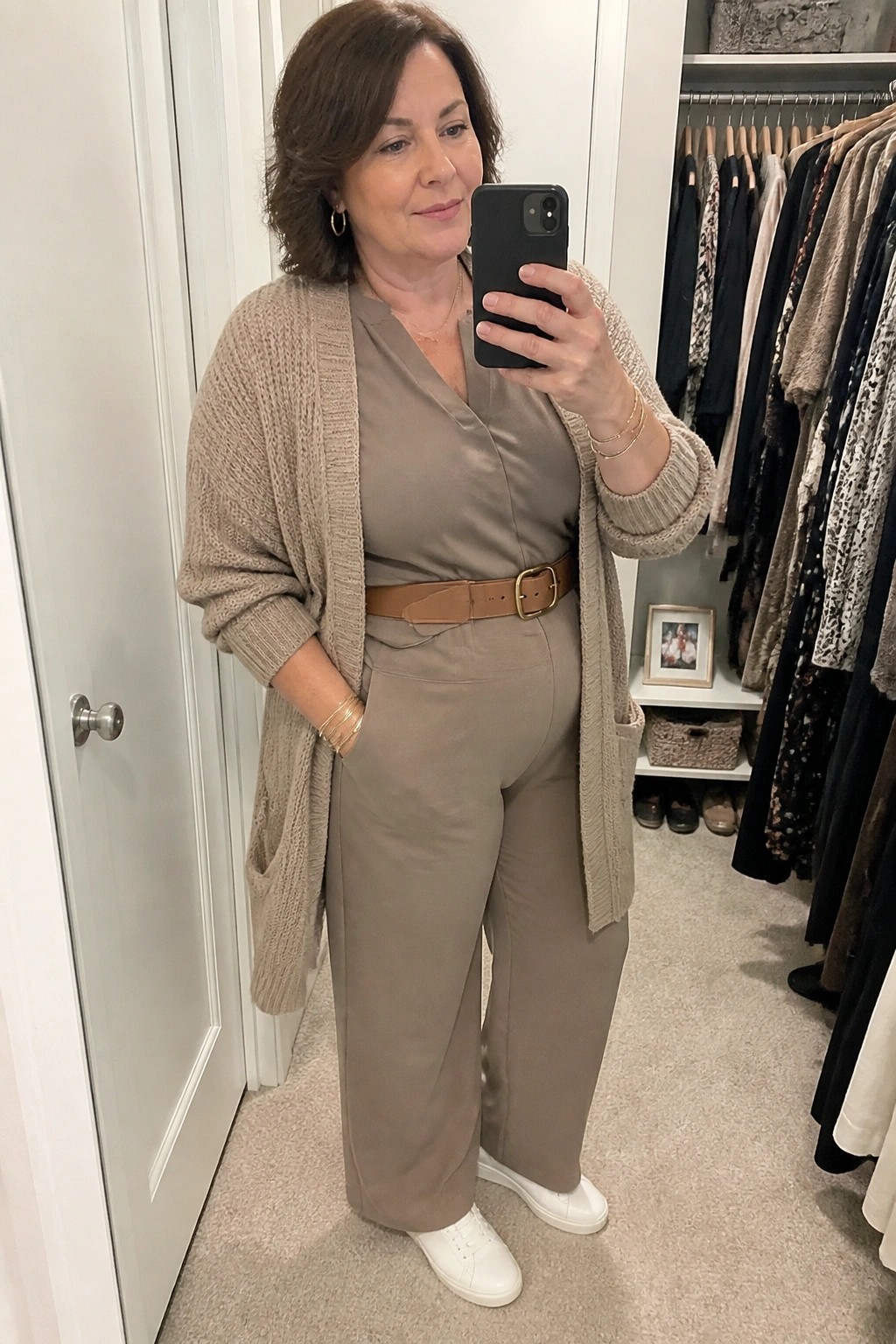Woman in taupe jumpsuit and beige cardigan taking mirror selfie in closet