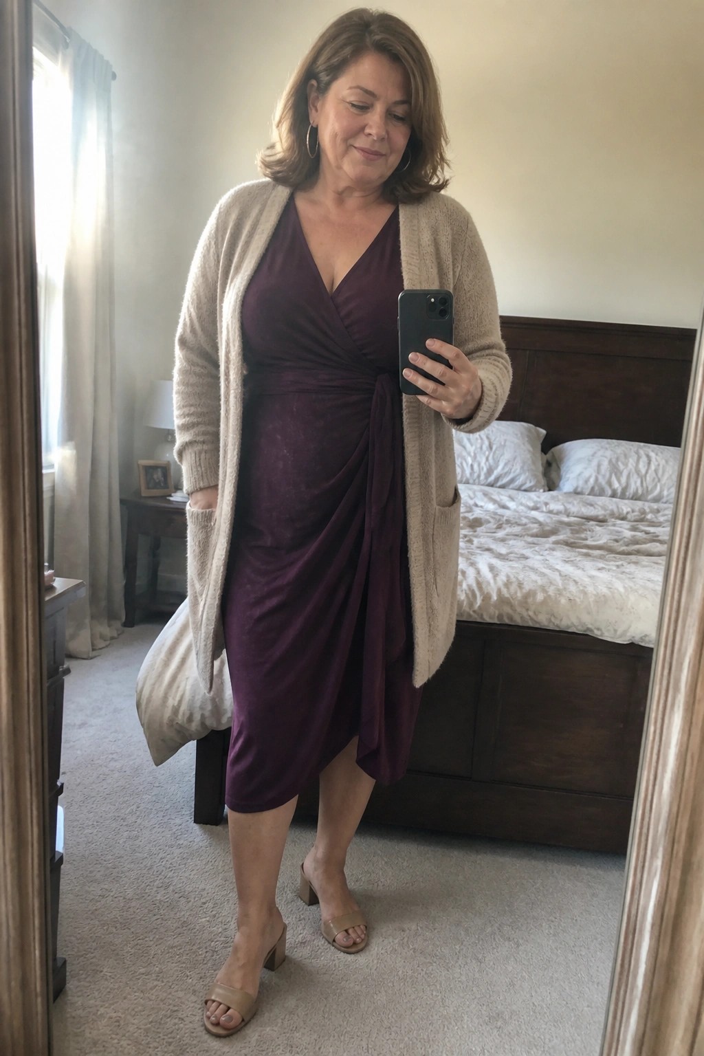 Woman in maroon wrap dress and beige cardigan taking bedroom mirror selfie.