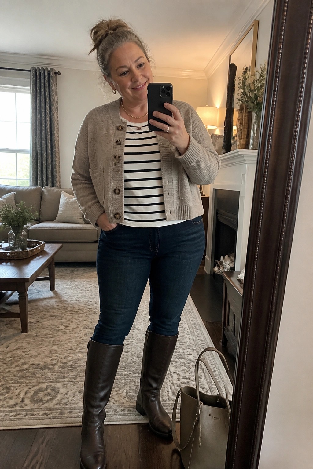 Woman over 50 wearing a beige button front cardigan over a black and white striped top paired with dark blue jeans and brown knee high boots while holding a phone in a living room setting.