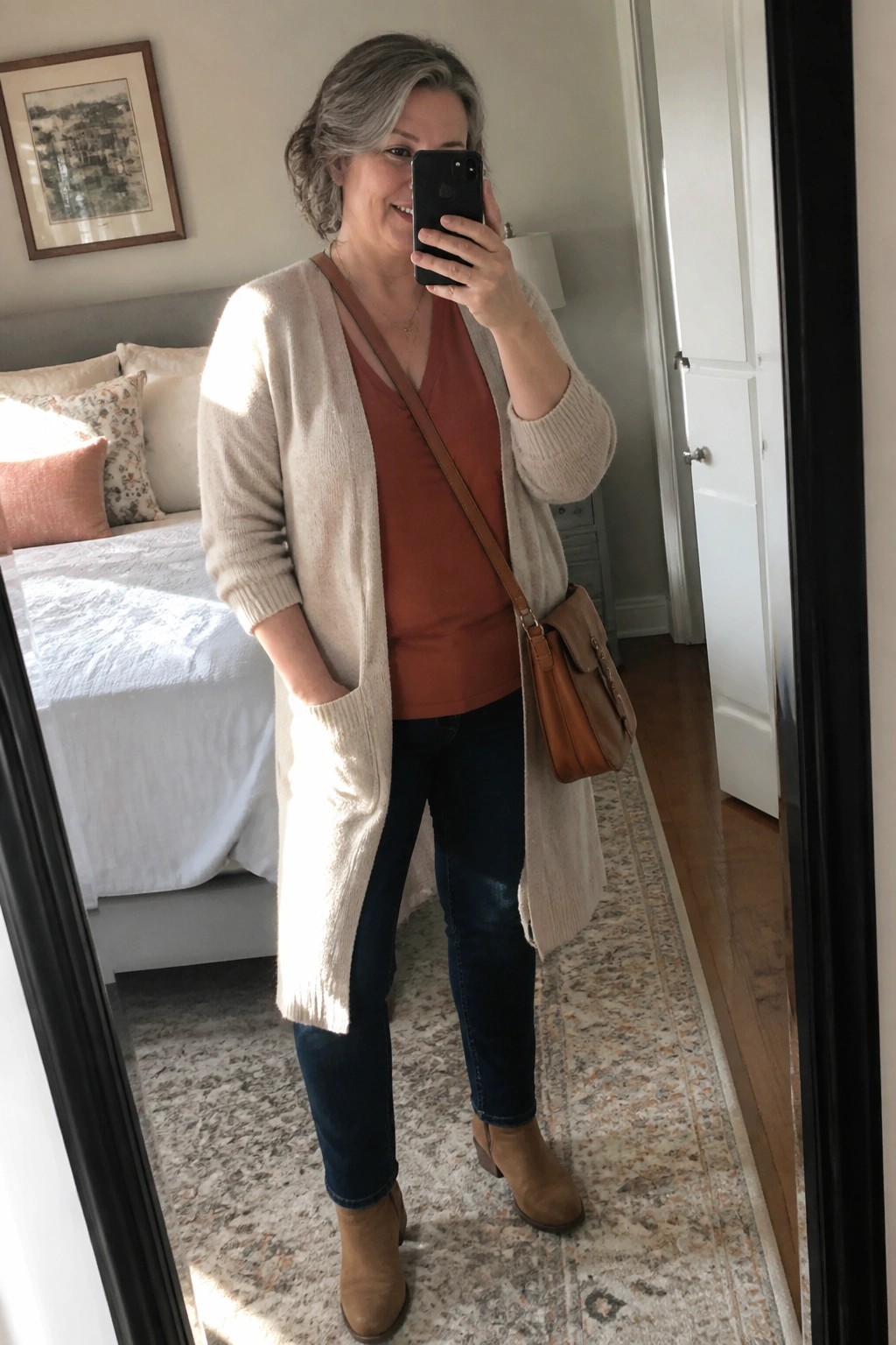 Woman in cream cardigan over rust top and jeans taking bedroom mirror selfie.