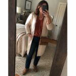 Woman in cream cardigan over rust top and jeans taking bedroom mirror selfie.