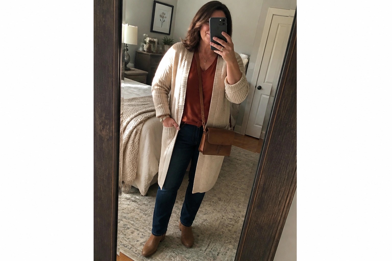 Woman in cream cardigan over rust top and jeans taking bedroom mirror selfie.