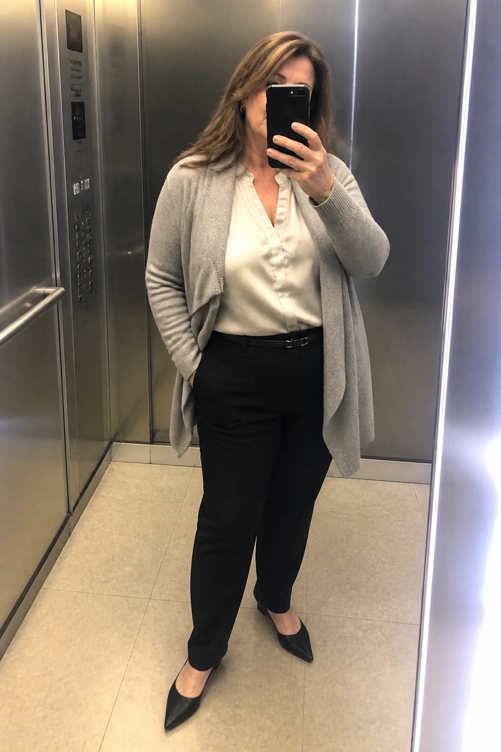 Woman in an elevator wearing an open gray cardigan over a white satin blouse tucked into black pants with a thin black belt and black pointed flats.