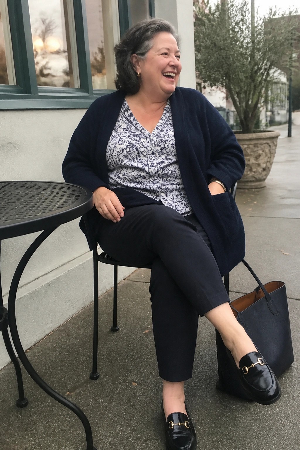 Smiling woman in navy cardigan over patterned blouse seated outdoors with black handbag