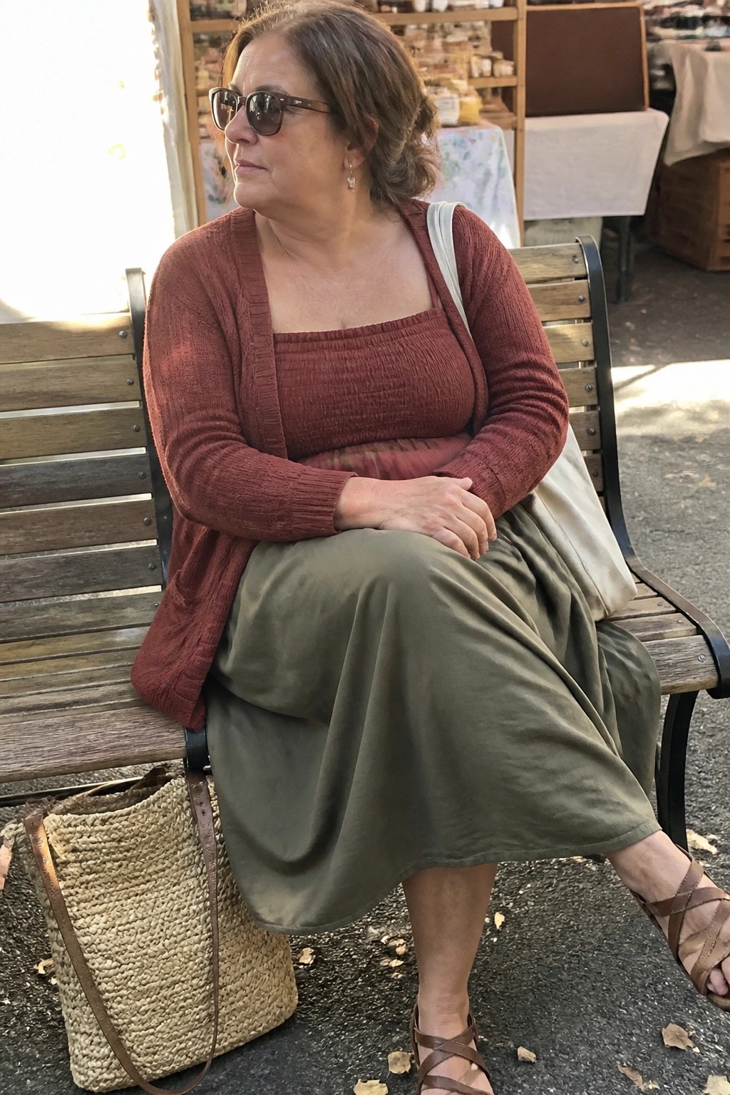 Woman in rust cardigan and olive skirt on wooden bench beside woven bag at market