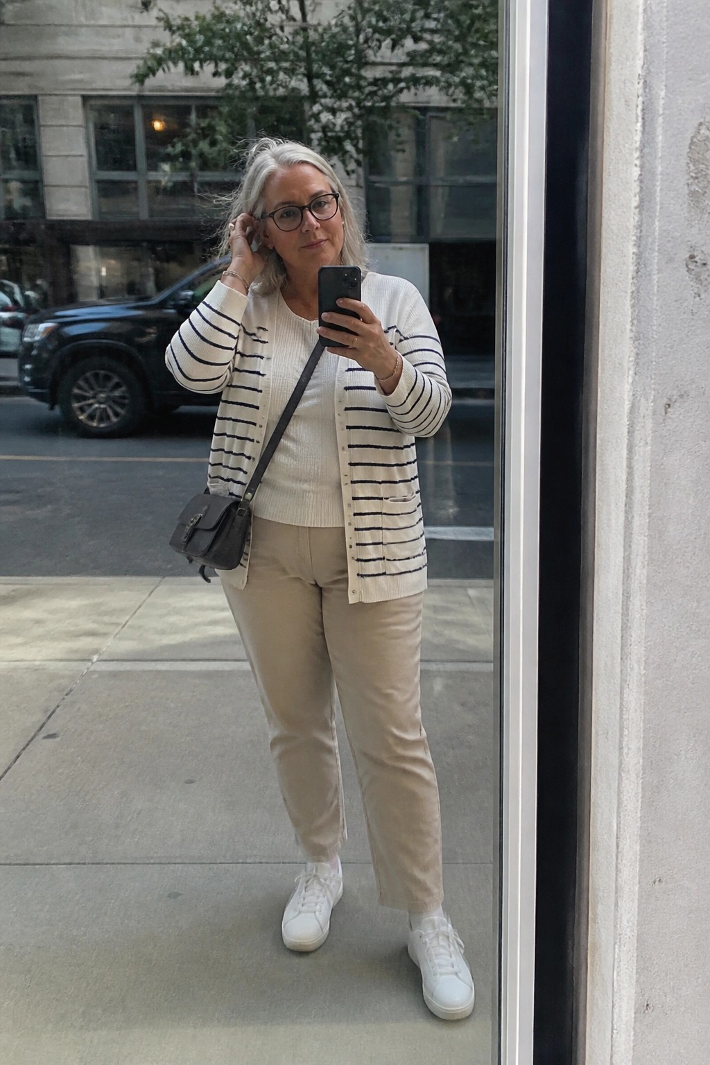 Woman over 50 wearing a navy striped button front cardigan over a cream knit top with beige straight leg pants white sneakers and a small black crossbody bag while taking a mirror selfie.