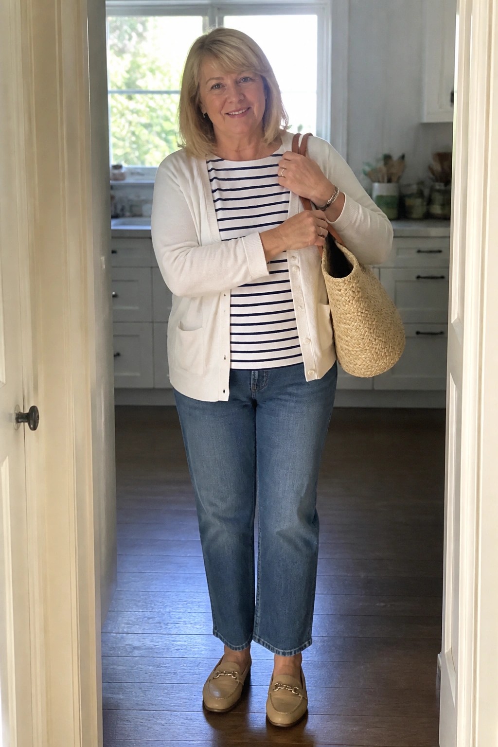 Woman in white cardigan over striped shirt with jeans holding woven straw bag in doorway.