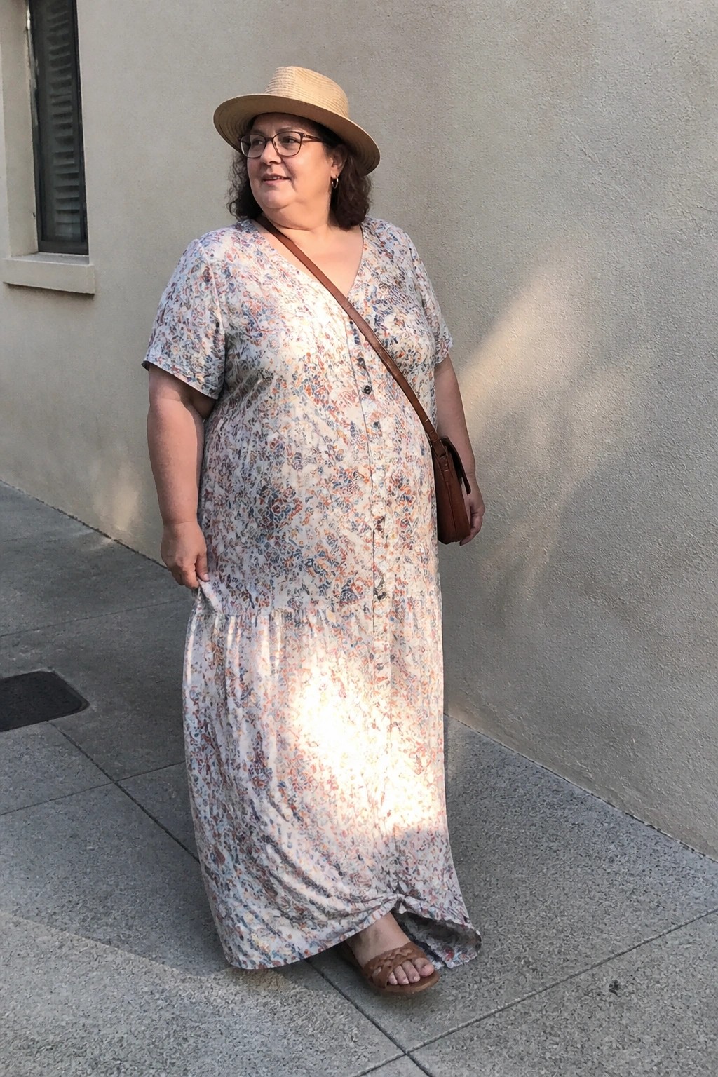 Plus size woman over 50 in a long floral button front maxi dress with short sleeves, straw hat, glasses, brown crossbody bag and woven sandals.