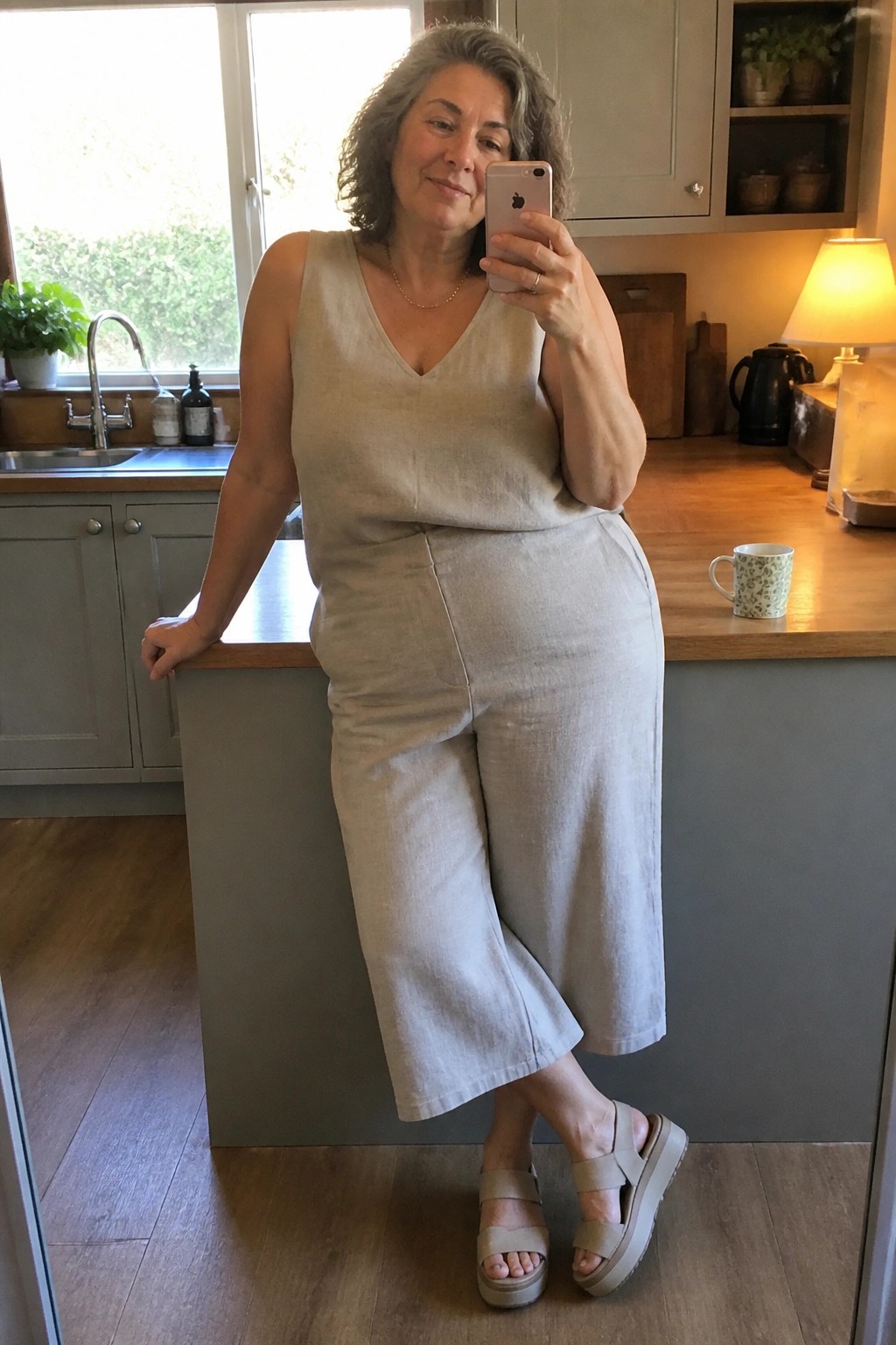Woman over 50 wearing a beige linen sleeveless V-neck tank top and matching wide-leg cropped pants with beige platform sandals, standing in a kitchen.