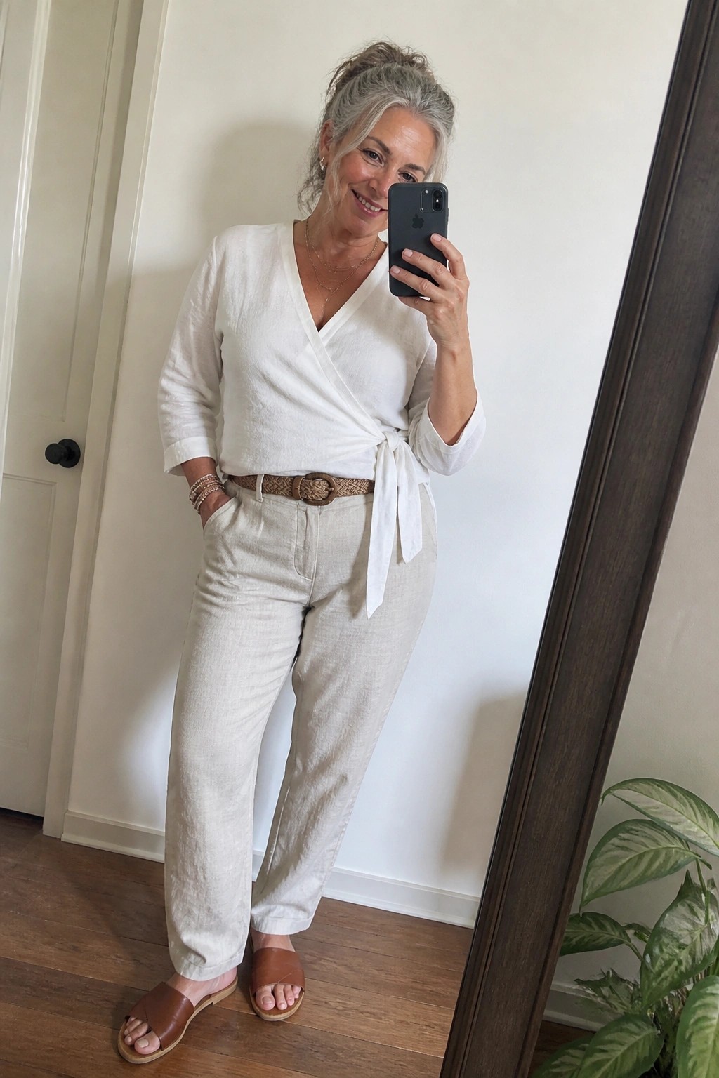 Smiling gray-haired woman in white wrap top and beige linen pants taking mirror selfie