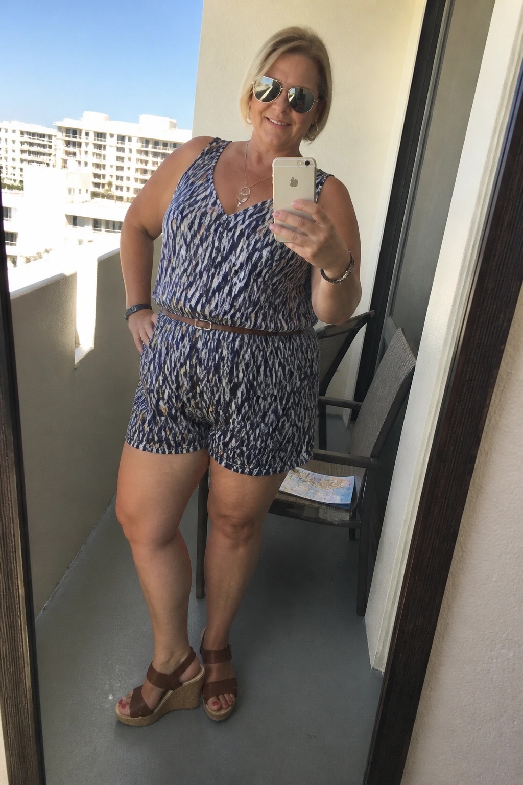 Woman in blue patterned romper taking selfie on sunny balcony with phone