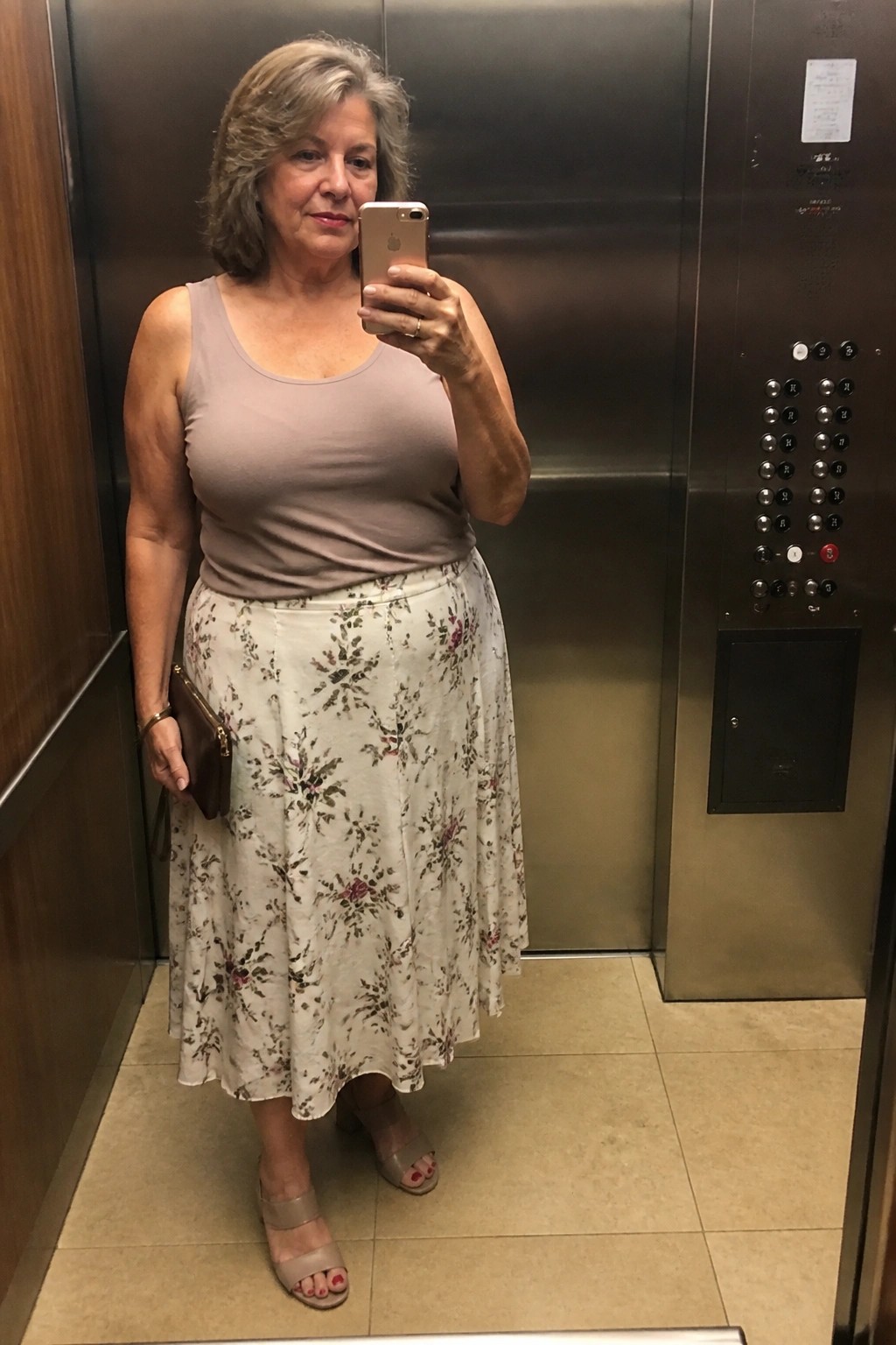 Woman with gray hair taking selfie in elevator wearing floral skirt and beige top