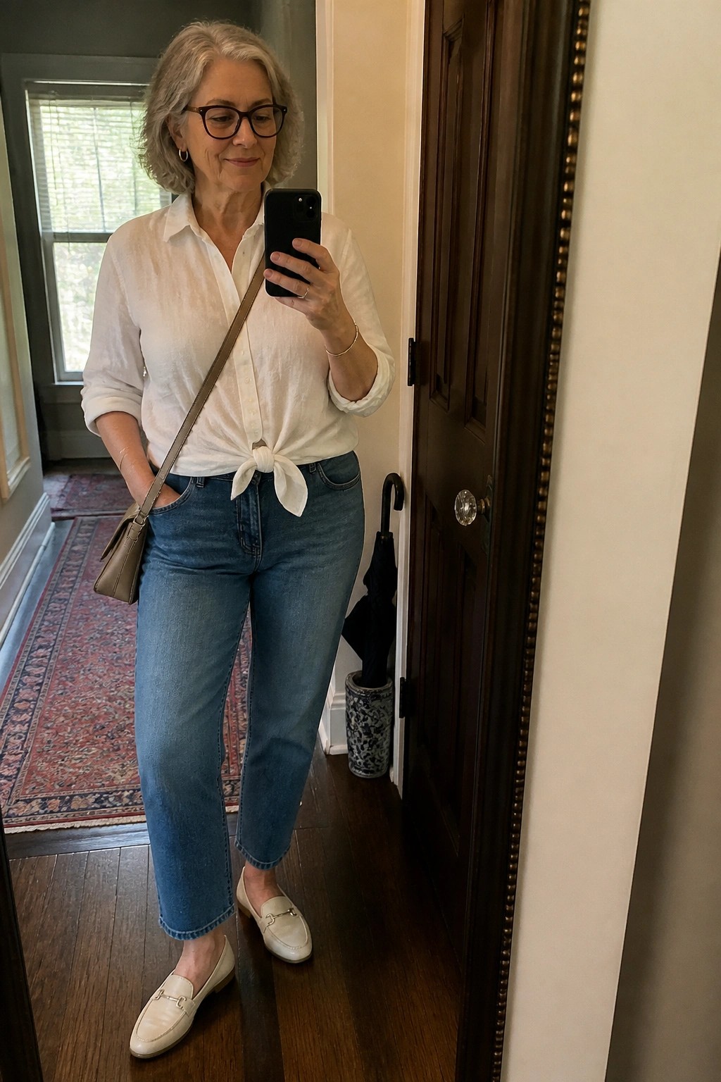 Gray-haired woman in tied white shirt and jeans taking mirror selfie with phone.