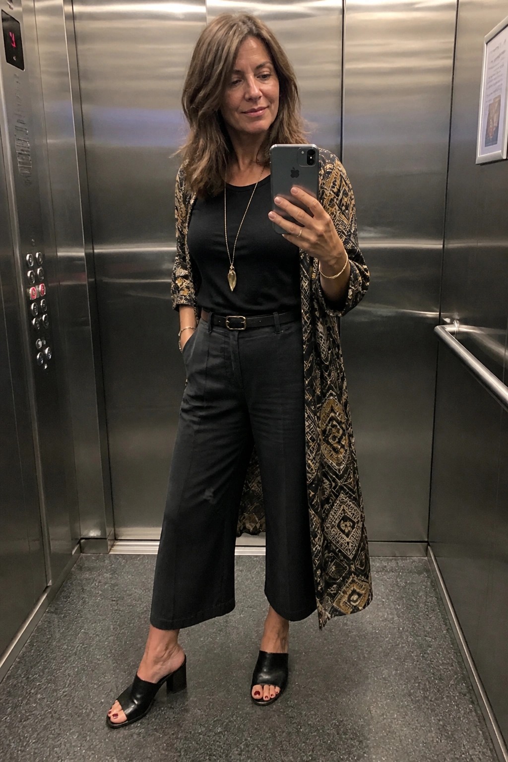 Woman in black top, wide-leg pants, and patterned jacket takes elevator selfie