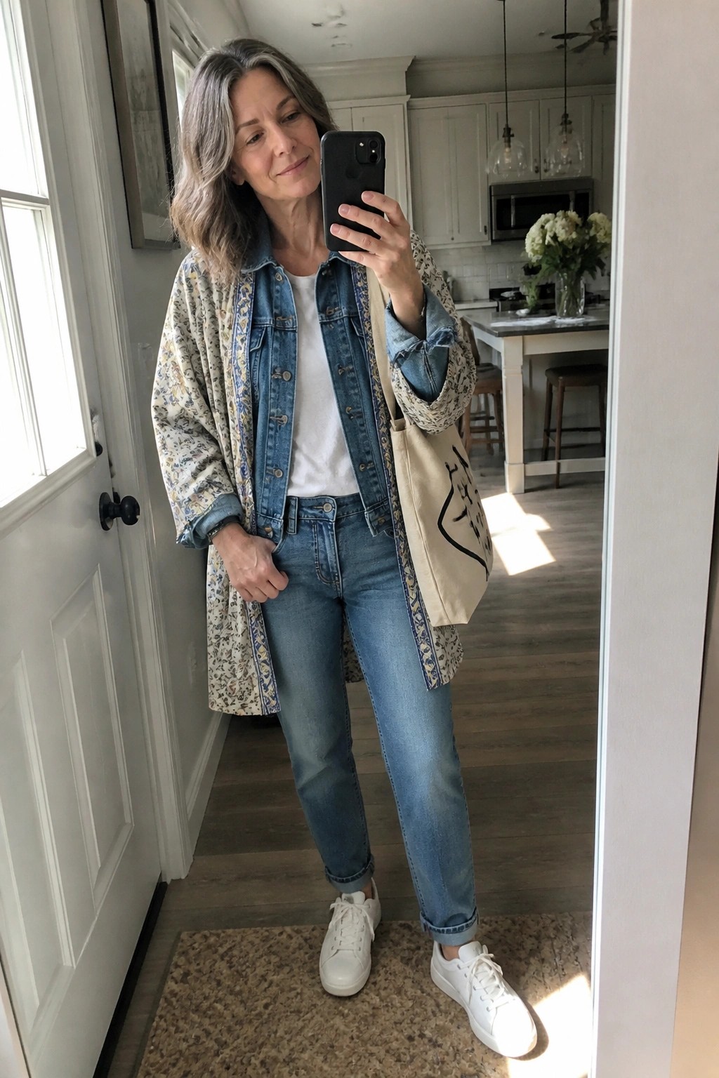Woman over 50 wearing a long floral print kimono jacket over a blue denim jacket, white t-shirt, blue jeans, and white sneakers while holding a beige tote bag in a bright kitchen setting.