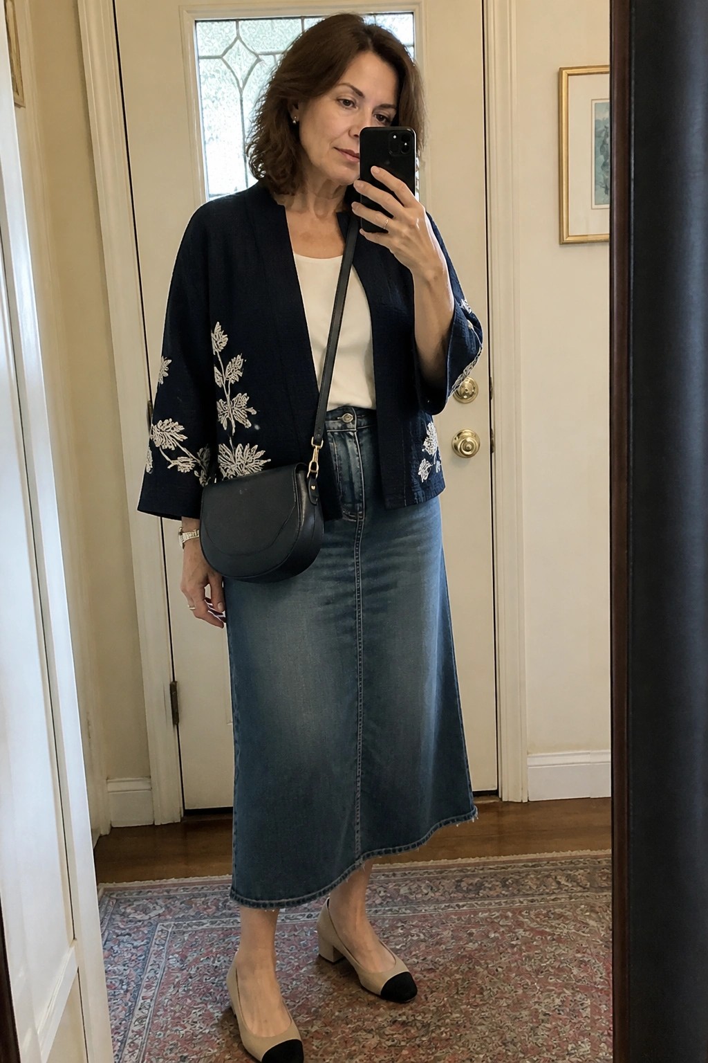 Woman in navy embroidered jacket and long denim skirt taking mirror selfie with phone
