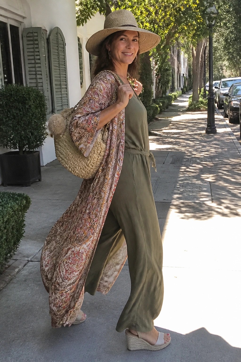 Woman in olive jumpsuit and floral kimono with straw hat carrying woven bag on sidewalk.