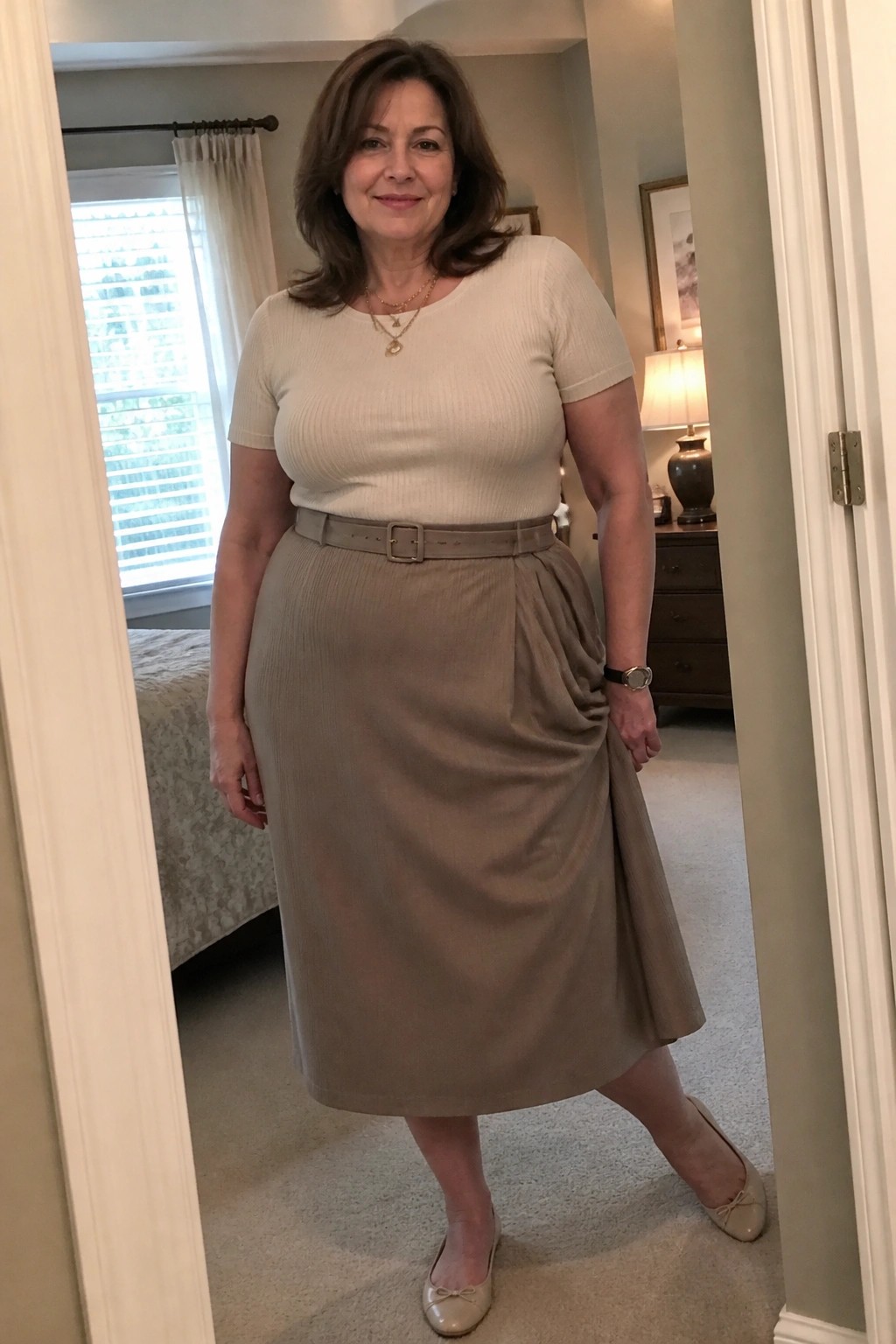Woman in beige top and belted taupe skirt standing in bedroom doorway