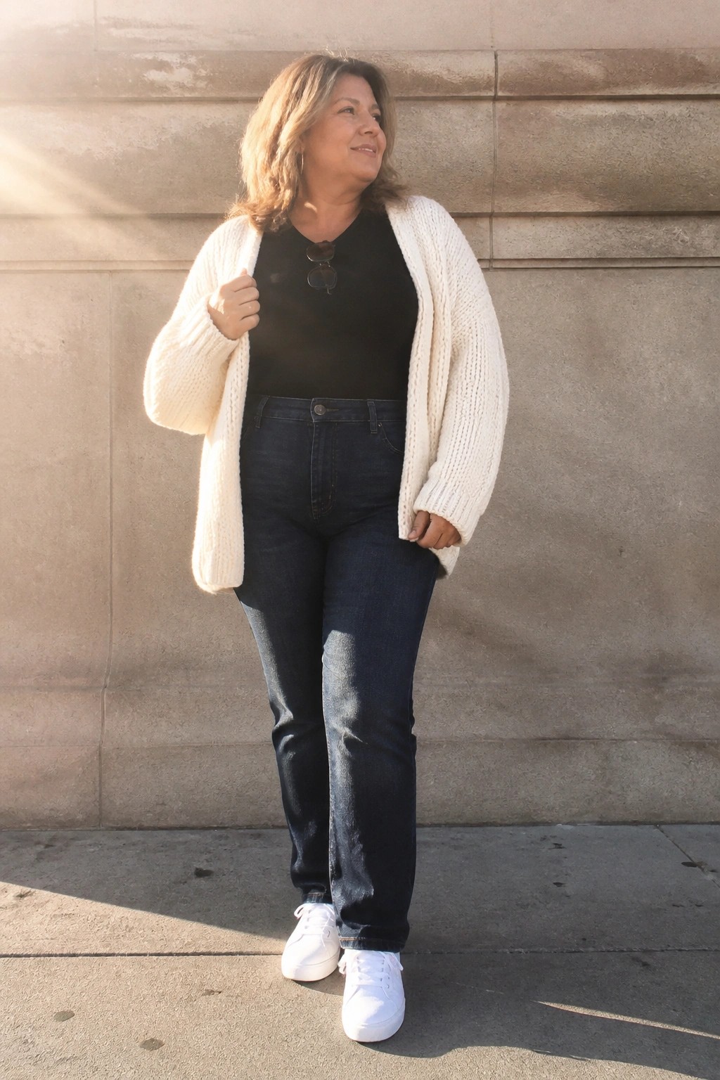 Woman in white knit cardigan and blue jeans by sunlit stone wall.