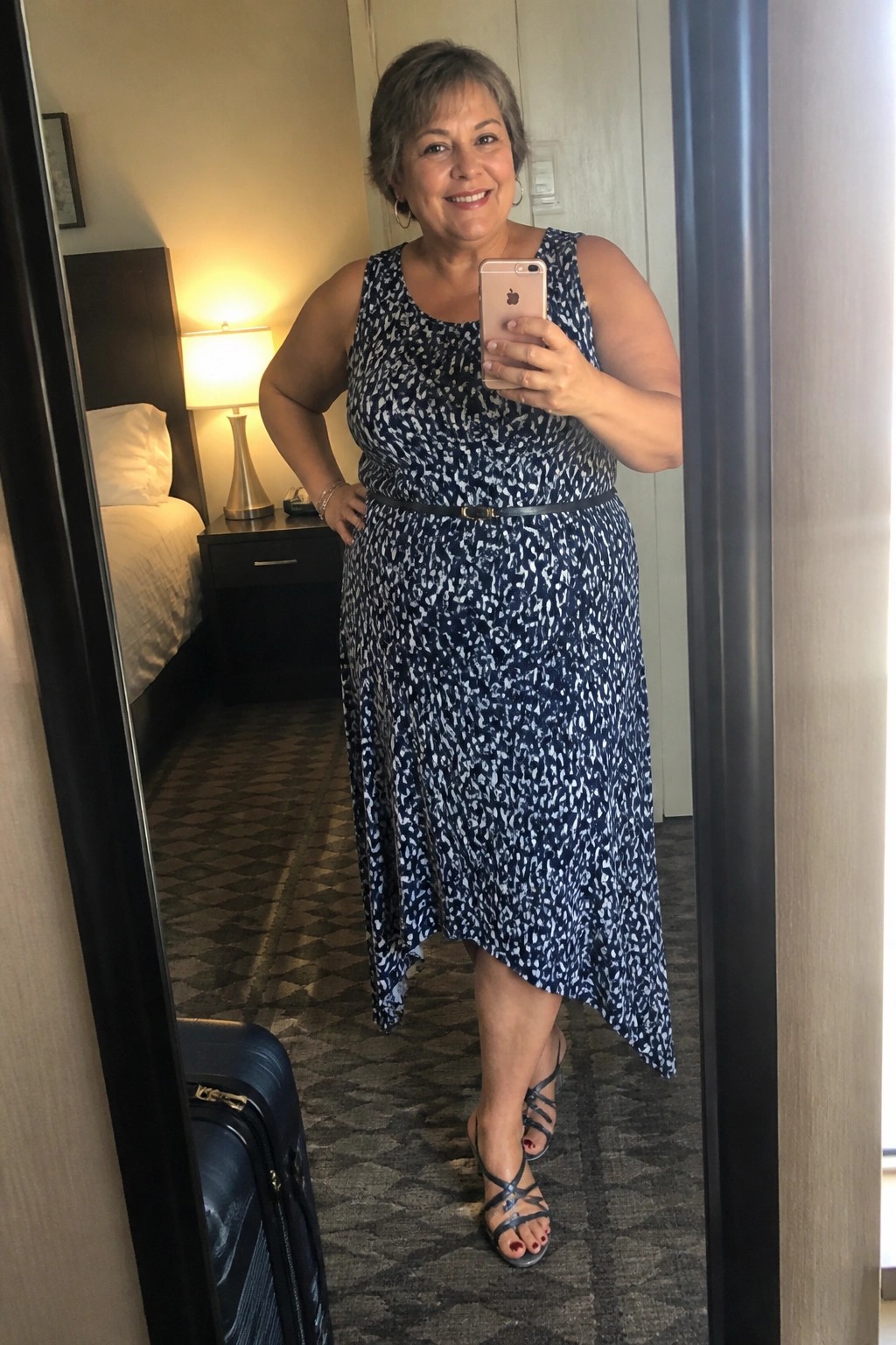 Smiling woman in blue patterned dress taking mirror selfie in hotel room