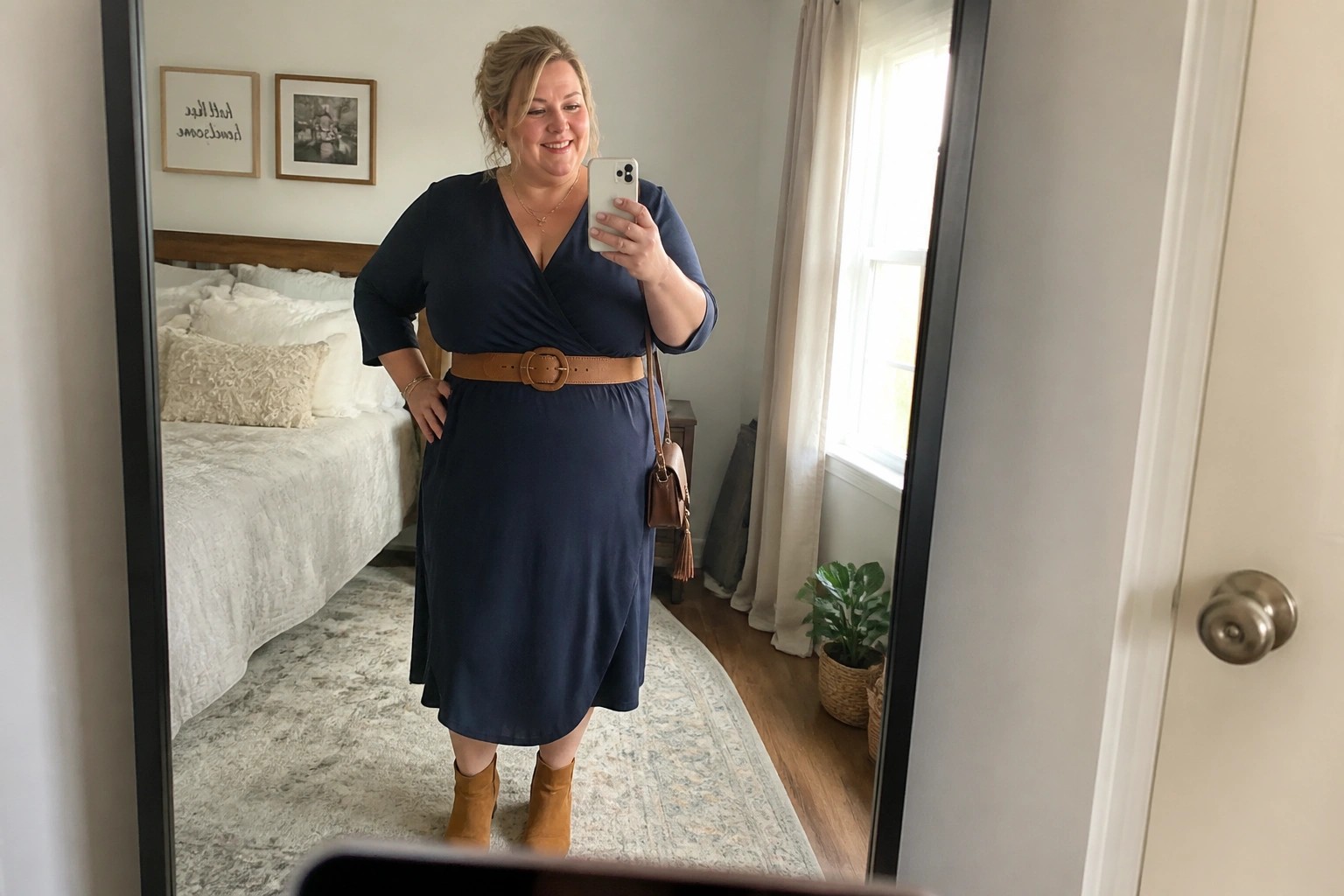 Woman in navy wrap dress with brown belt taking bedroom mirror selfie