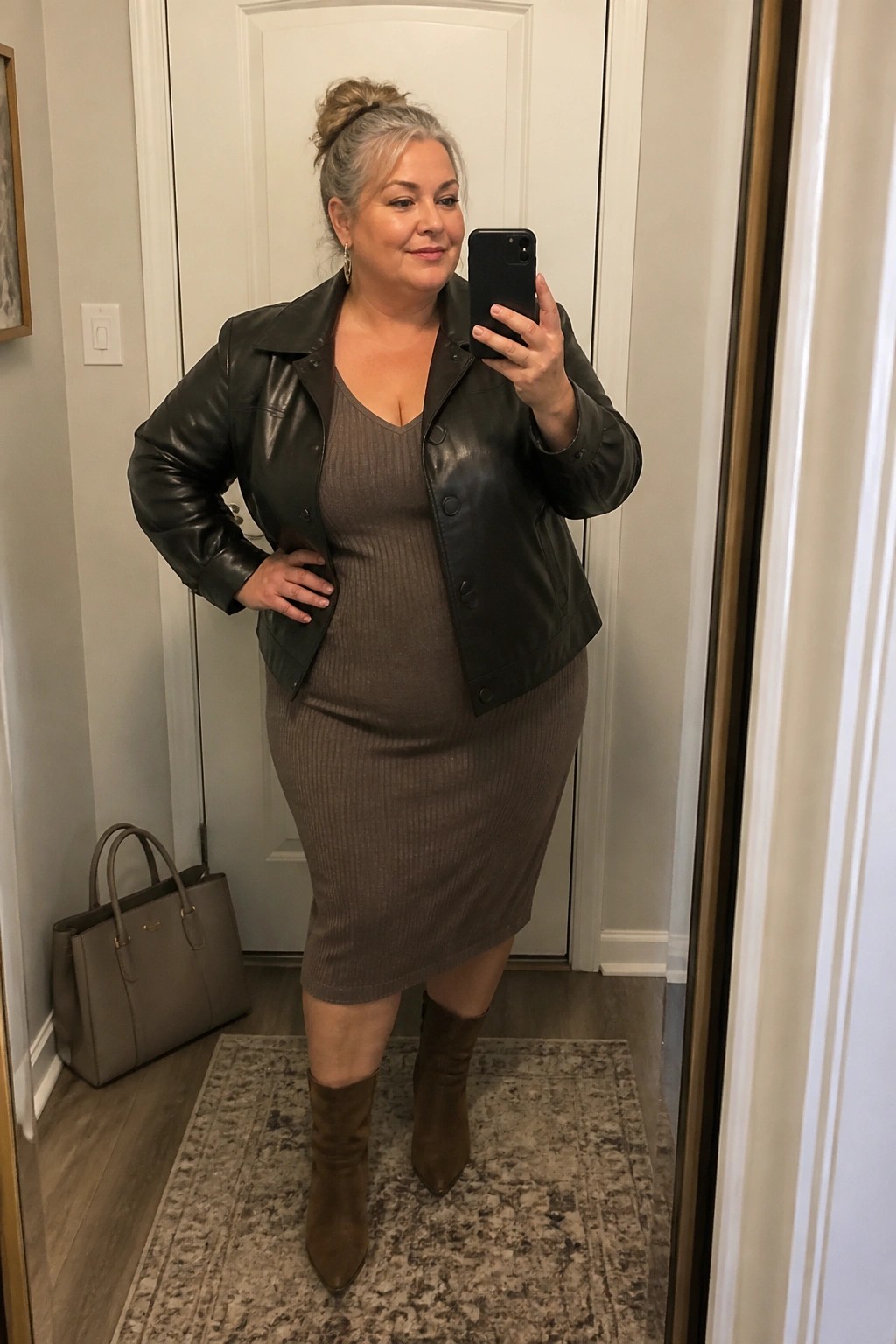 Gray-haired woman in brown ribbed dress and black leather jacket taking mirror selfie.
