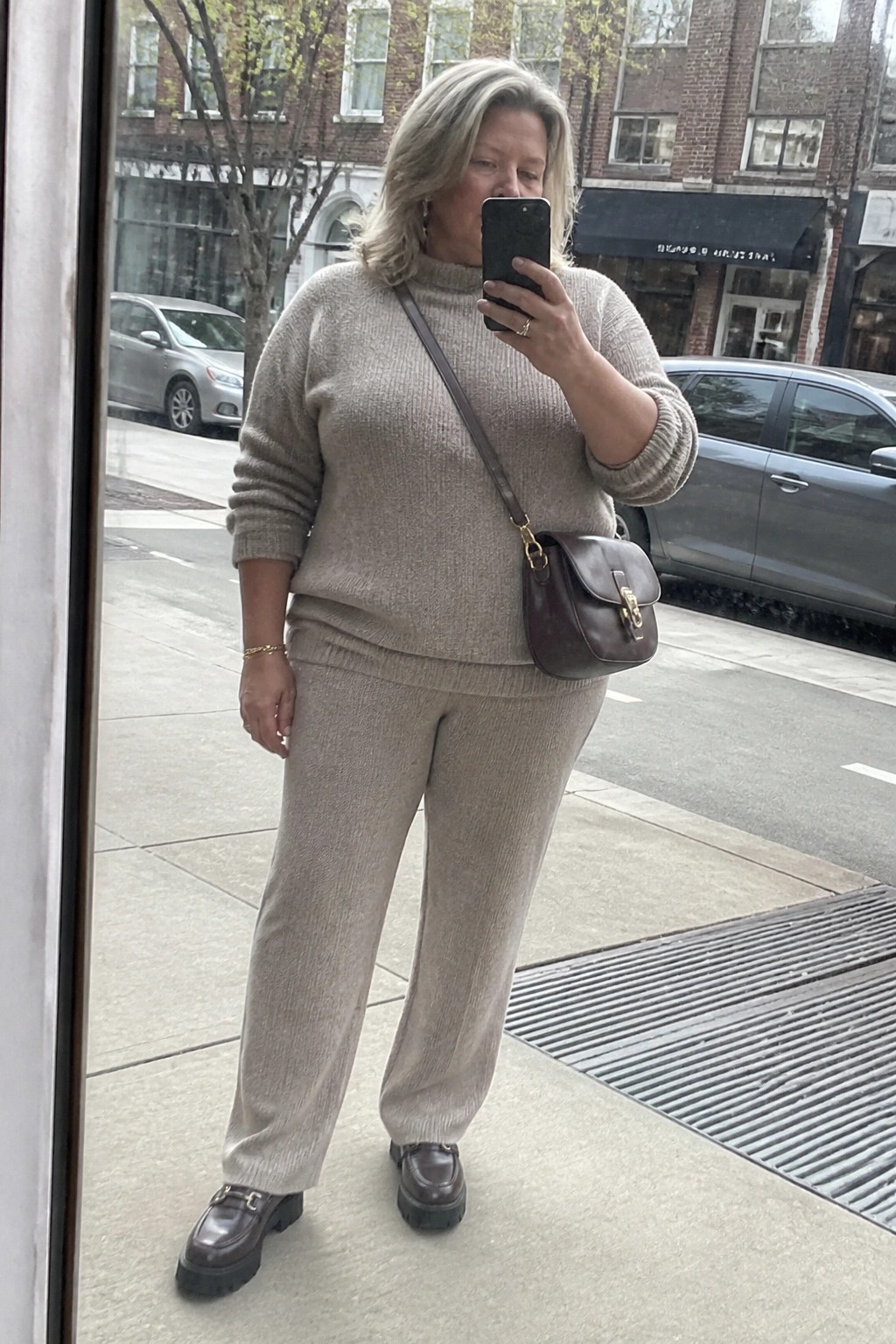 A plus size woman over 50 wearing a beige matching knit sweater and pants set with brown loafers and a brown leather crossbody bag taking a mirror selfie outdoors.