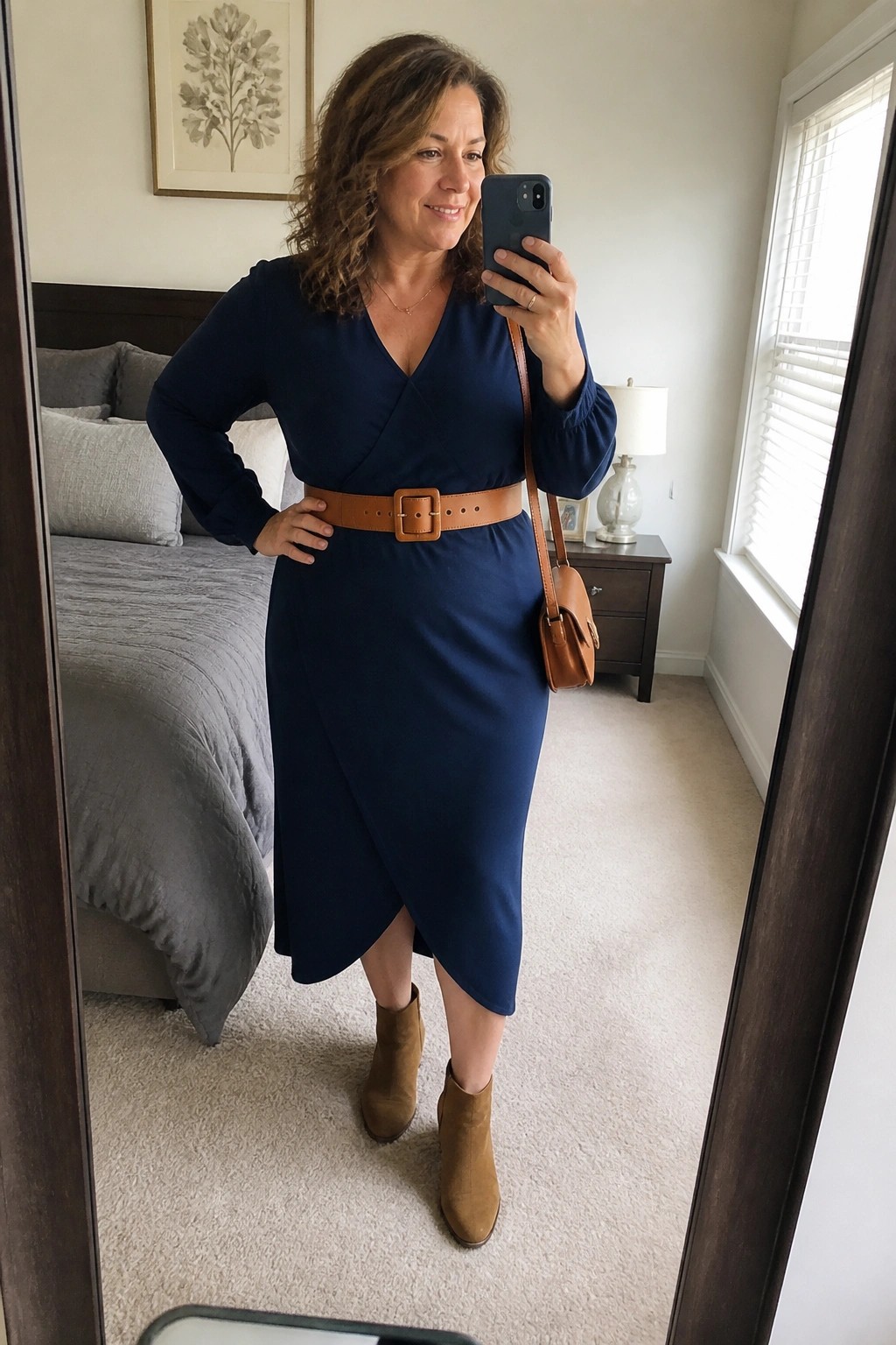 Woman in navy wrap dress with brown belt taking bedroom mirror selfie