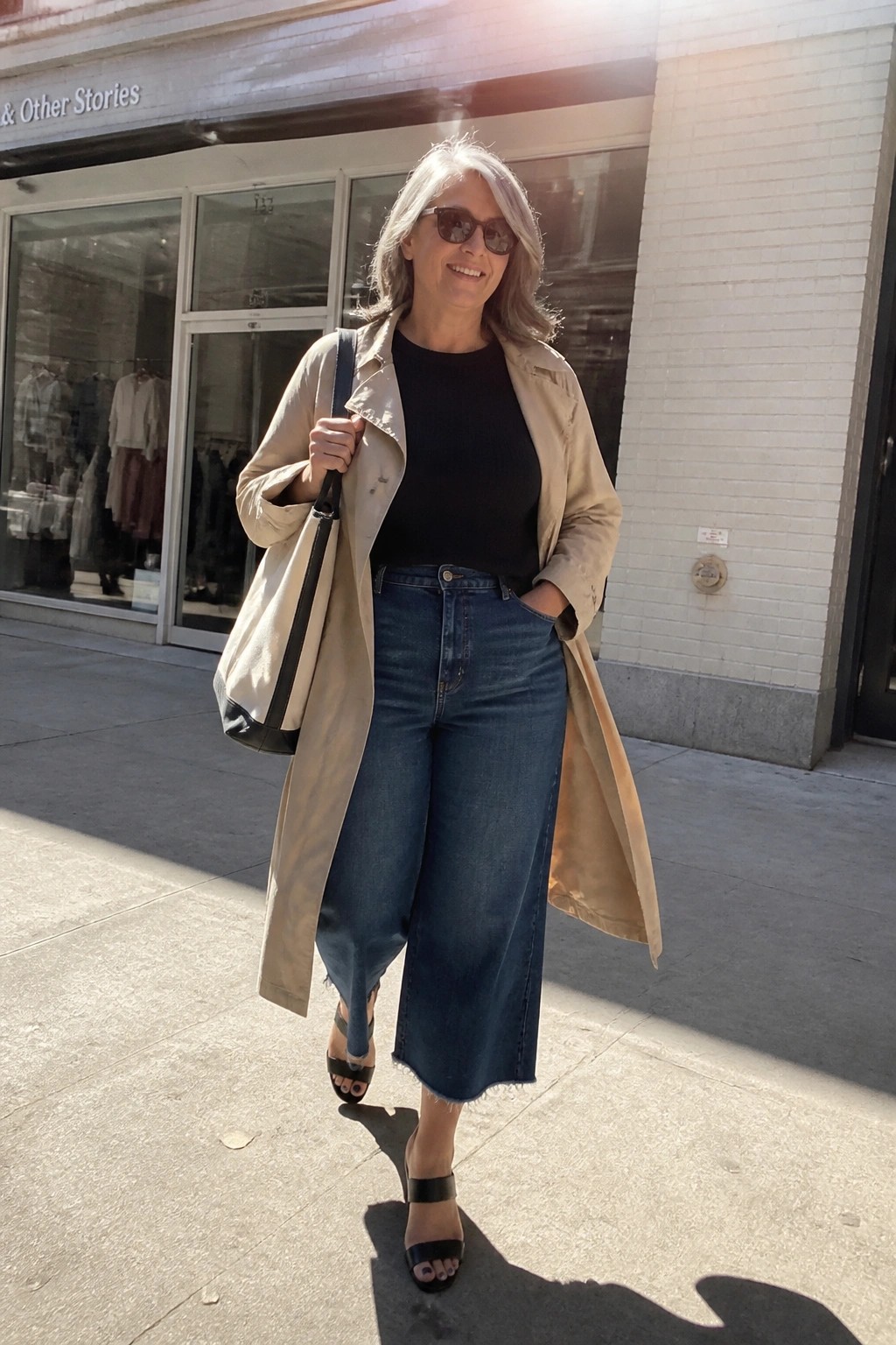 Smiling gray-haired woman in beige trench coat, black top, and blue wide-leg jeans on sunny sidewalk