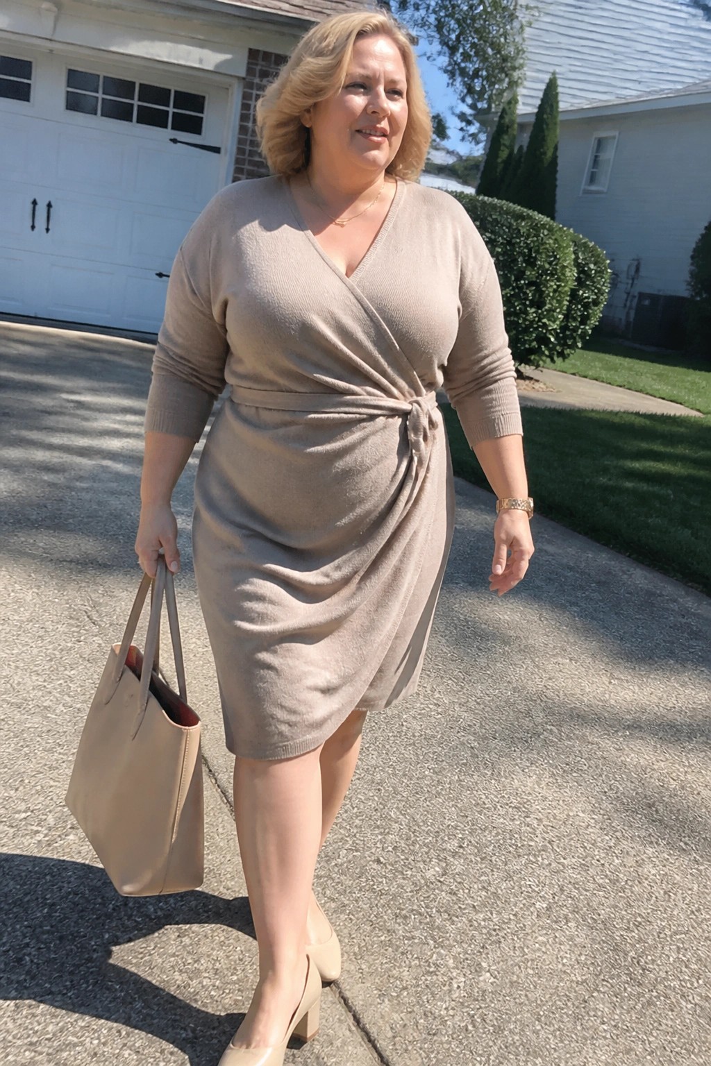 Plus-size blonde woman in beige wrap sweater dress holding tote bag on driveway