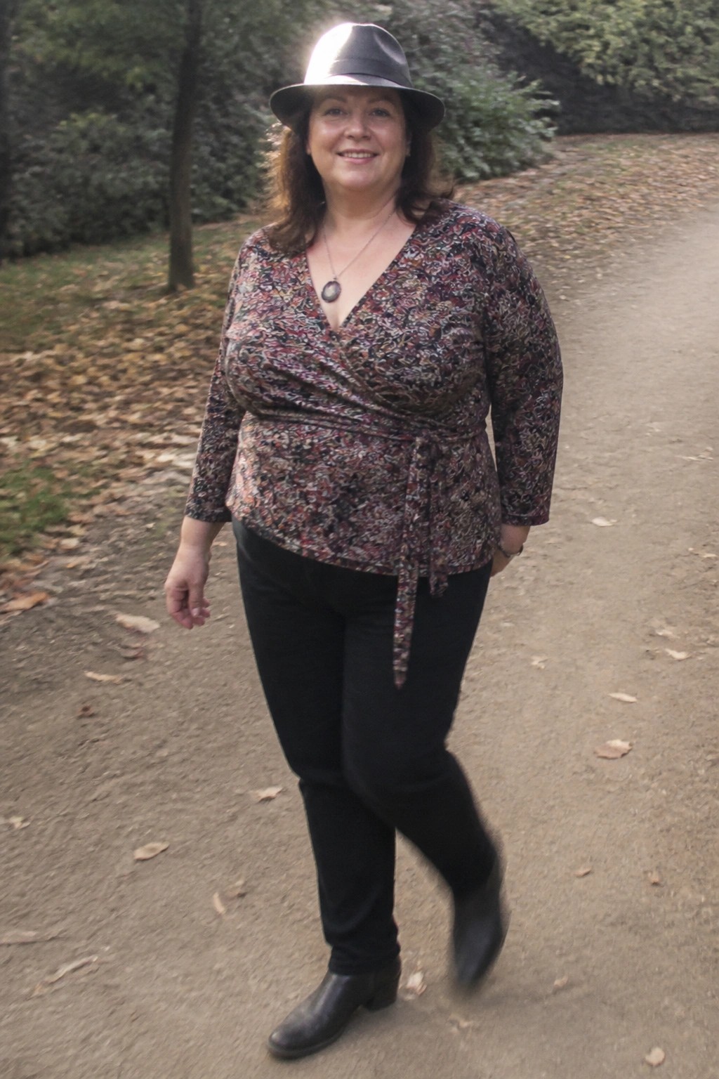 Woman in floral wrap top, black hat, pants and boots on leafy path
