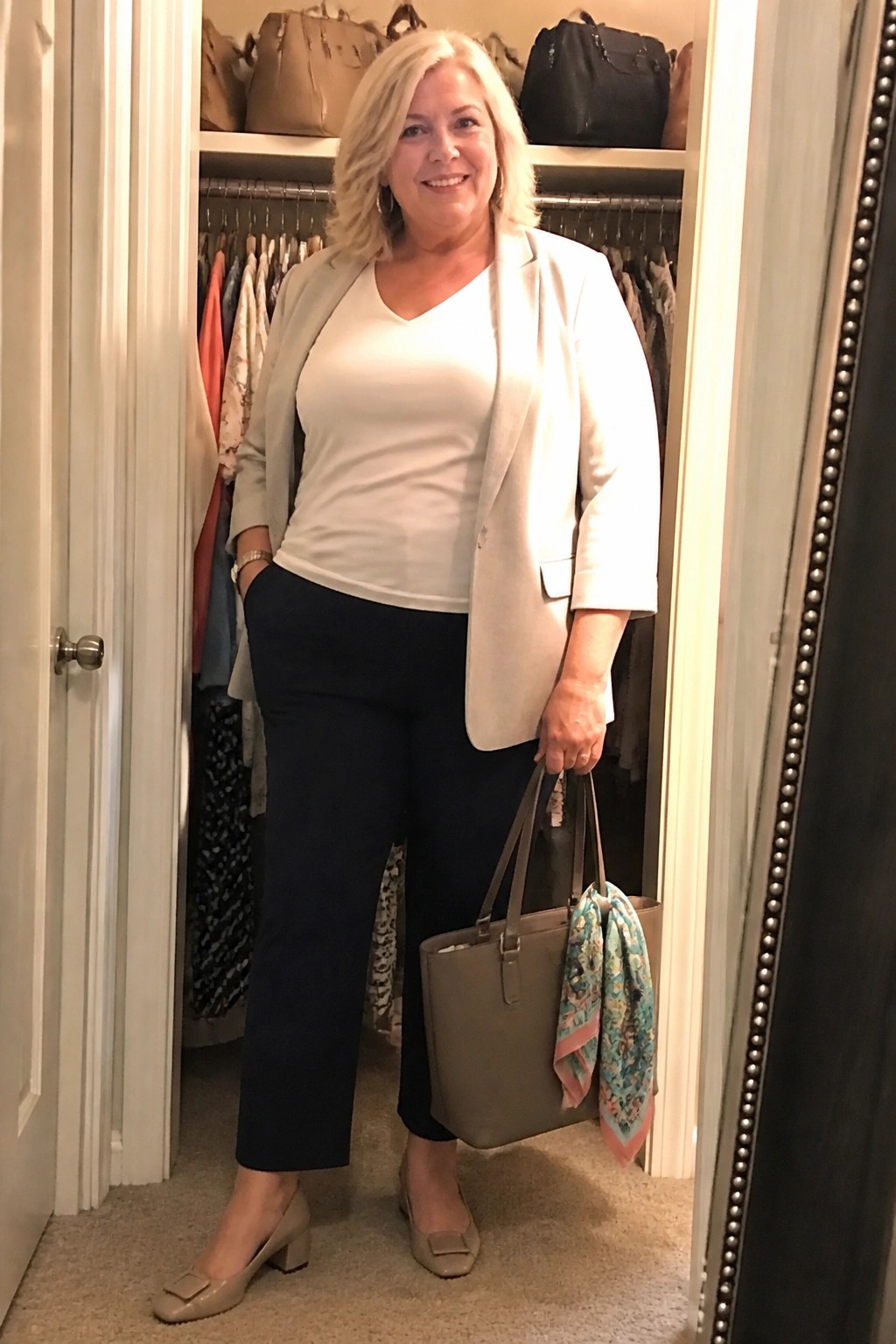 Smiling blonde woman in beige blazer and navy pants holds handbag in closet.