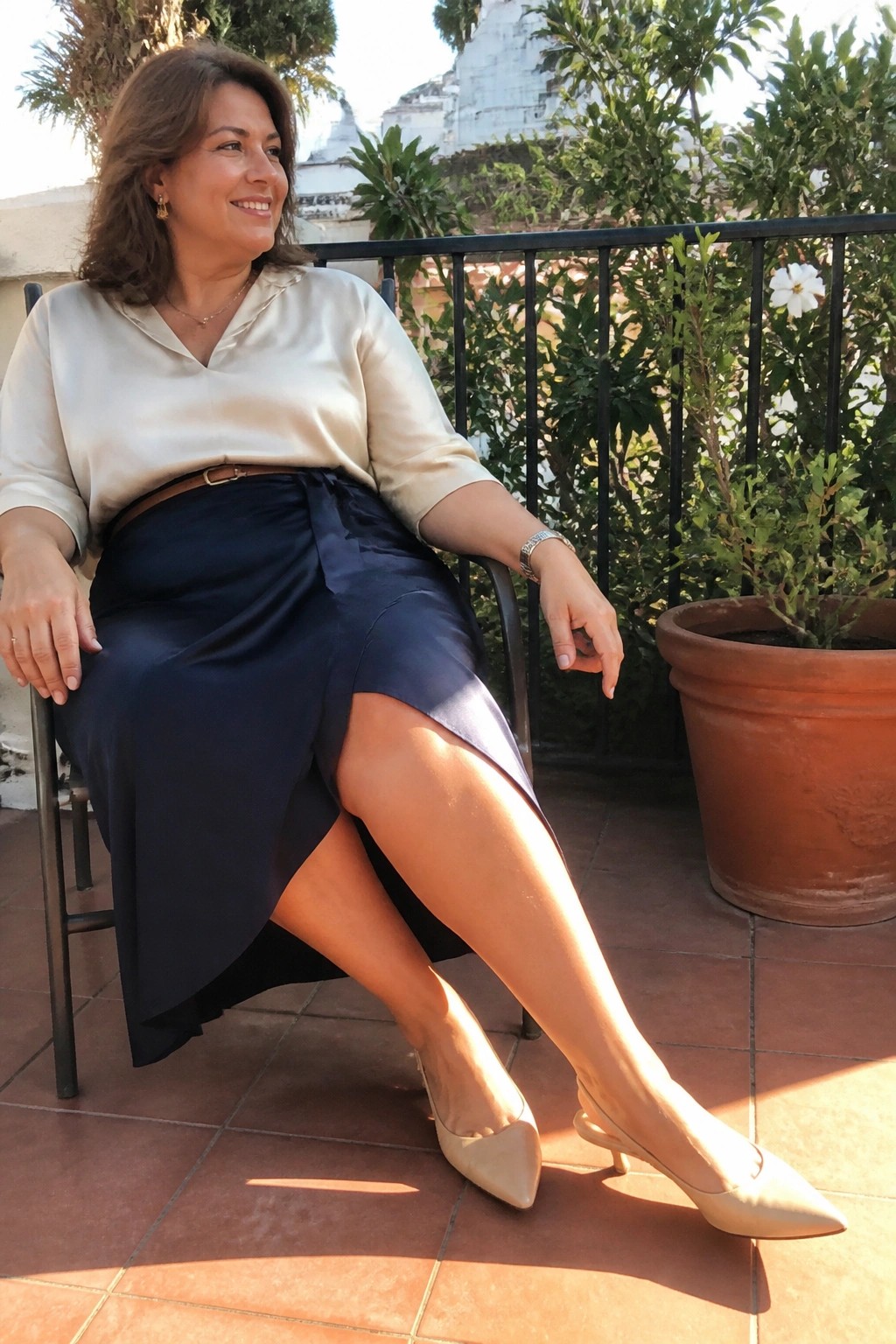 Woman in beige satin blouse and navy wrap skirt on sunny balcony with plants.