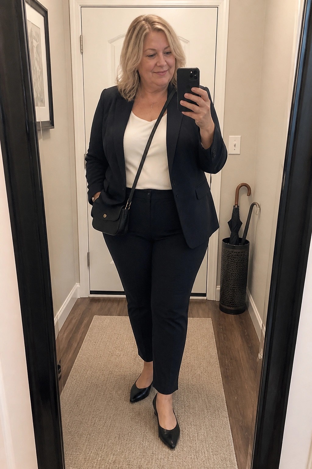 Blonde woman in black blazer and pants taking mirror selfie in hallway