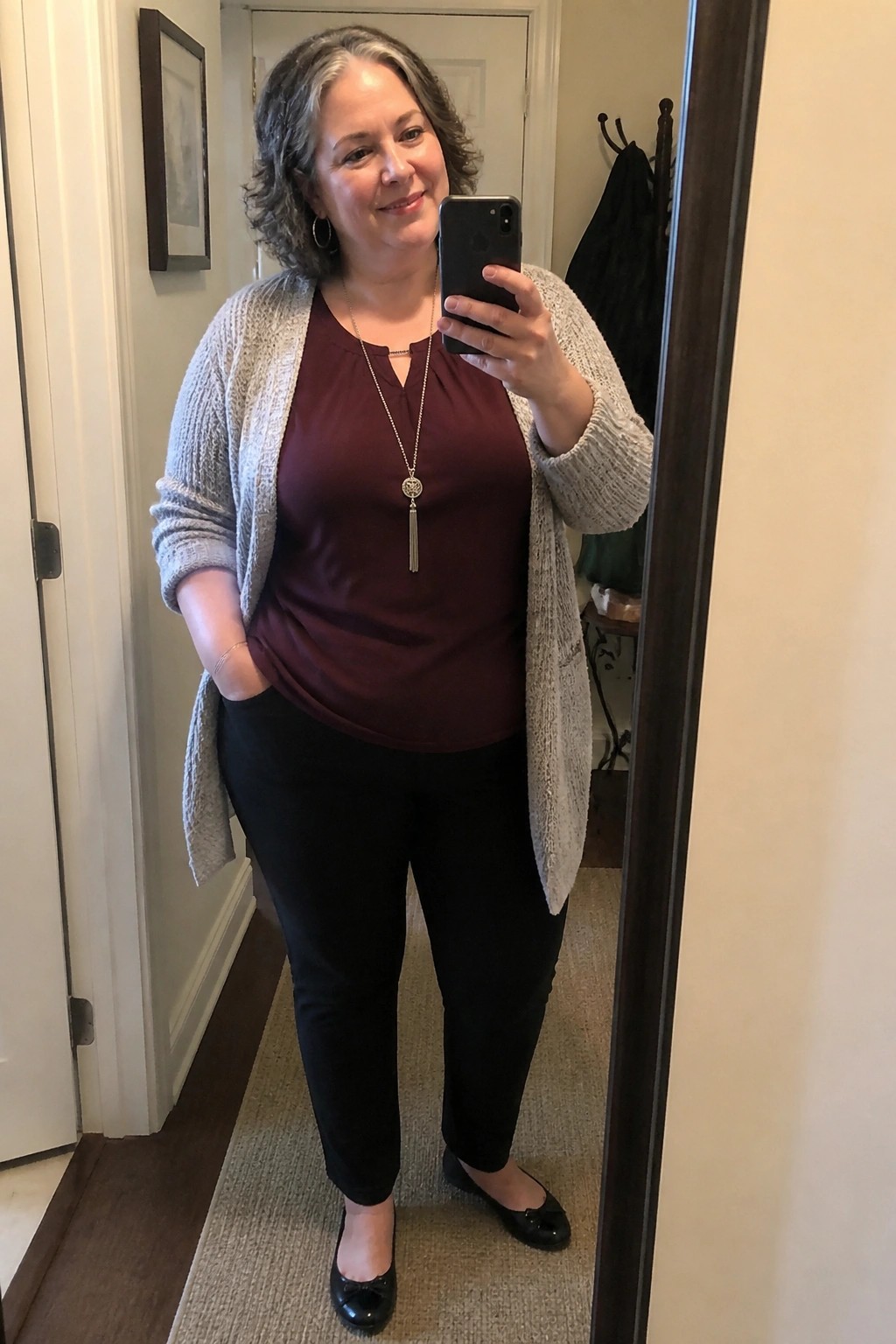Woman with curly gray hair in maroon top and gray cardigan taking mirror selfie