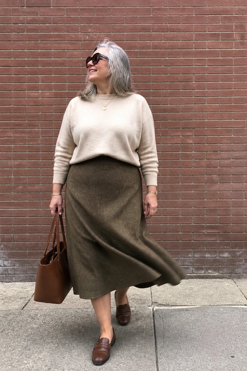 Smiling gray-haired woman in beige sweater and flowing olive skirt with brown bag