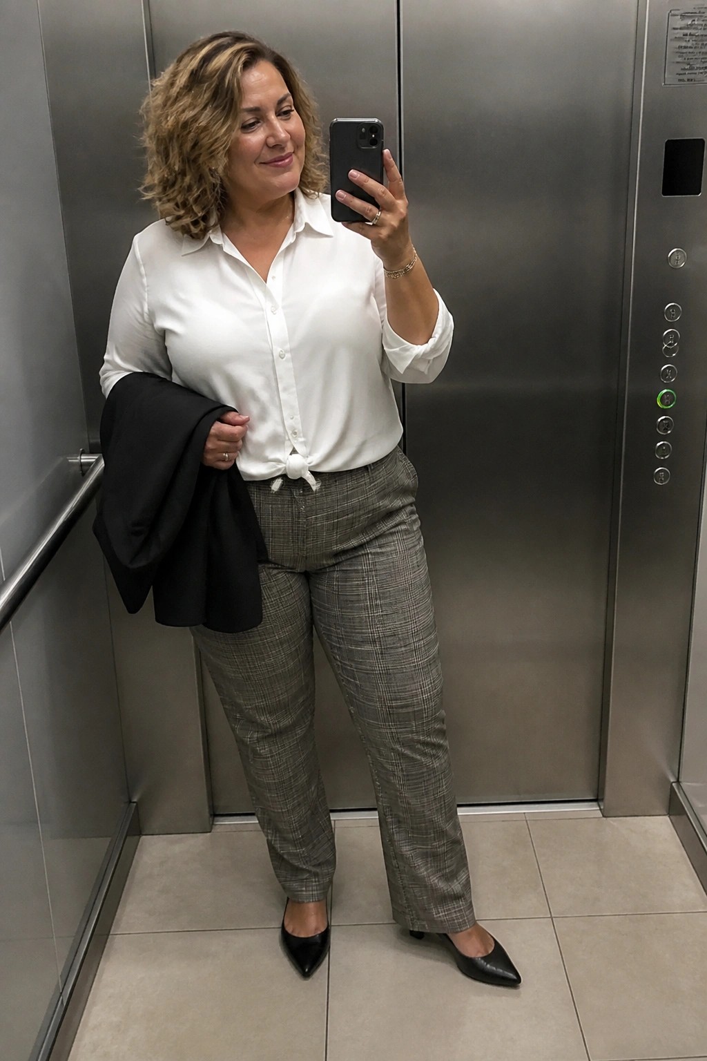 Woman in white shirt and plaid pants taking selfie in elevator holding black jacket
